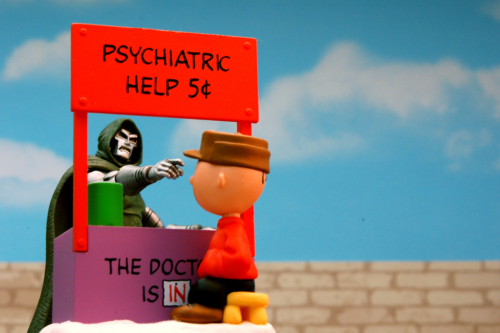 Comic book villain raising a finger to another toy character, under a booth labeled "psychiatric help 5 cents"