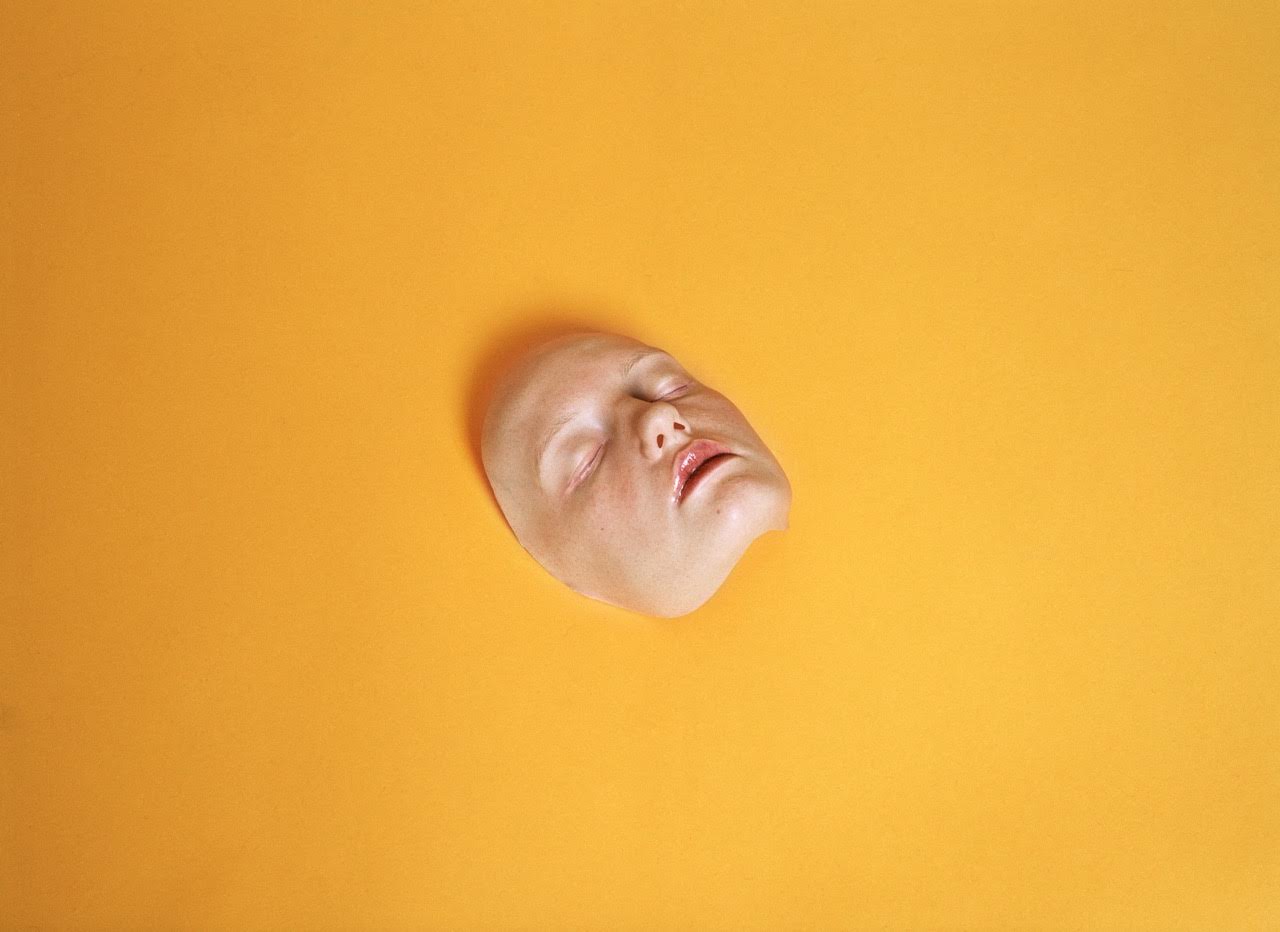 A person's face, white, with closed eyes and slightly open mouth, surrounded entirely by a dark yellow