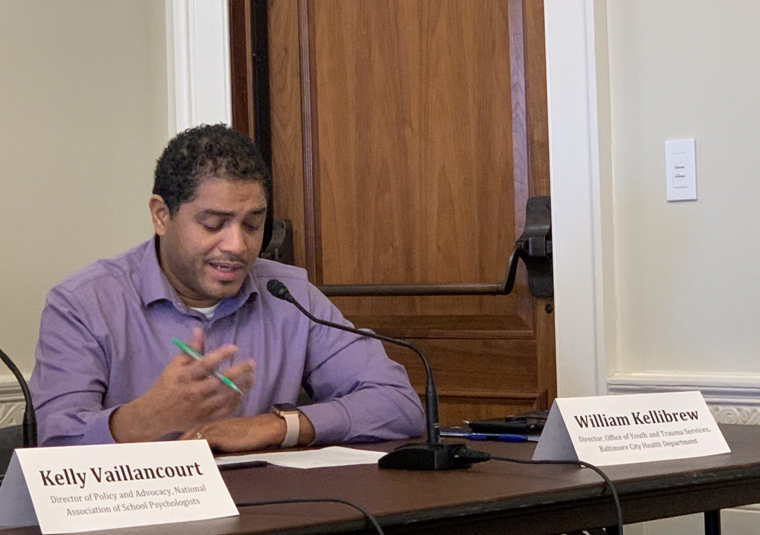 William wears a lavender shirt and speaks into a table microphone.