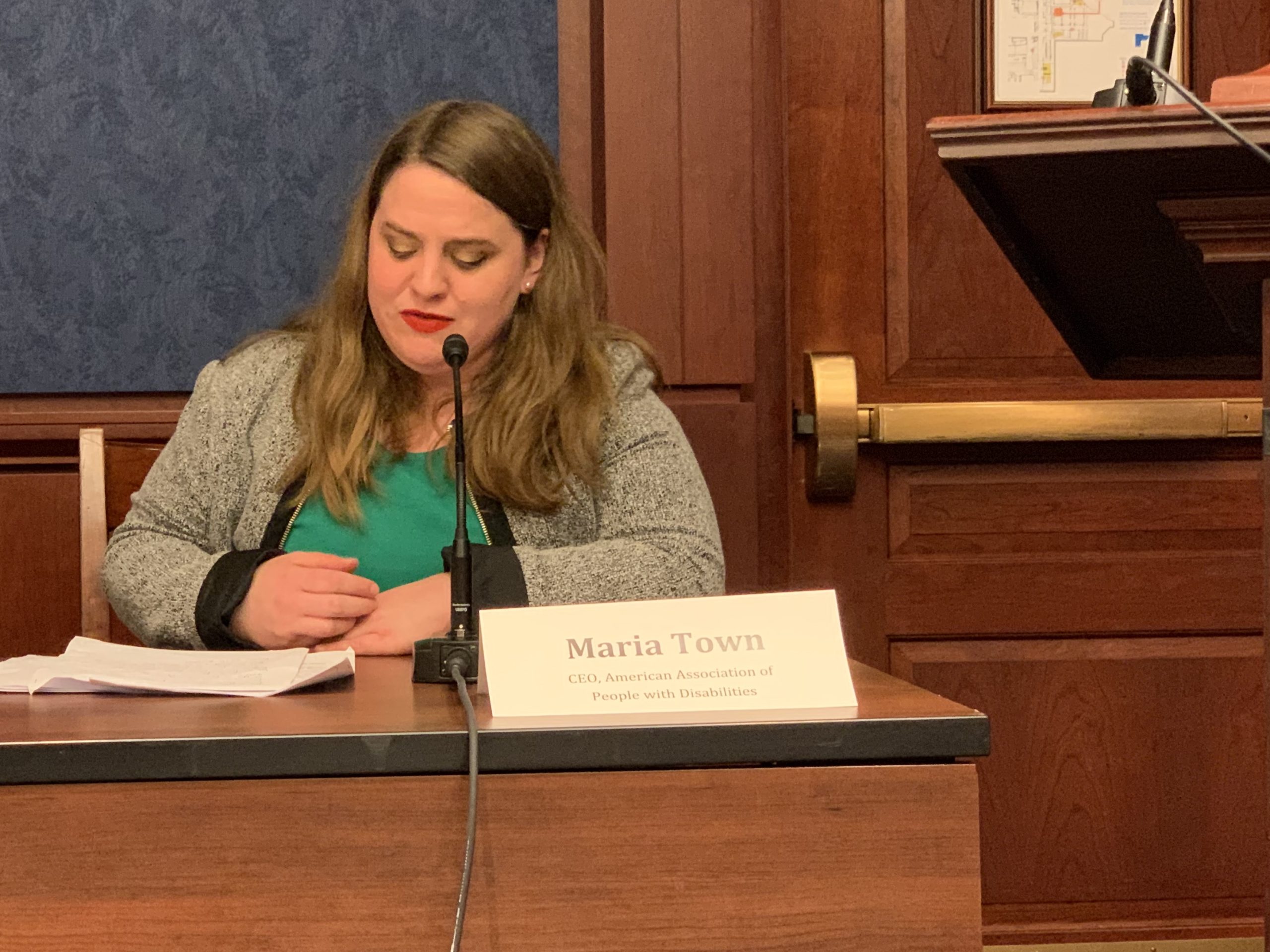 Maria Town, wearing a green shirt and grey sweater, sits as a table and speaks into a microphone.