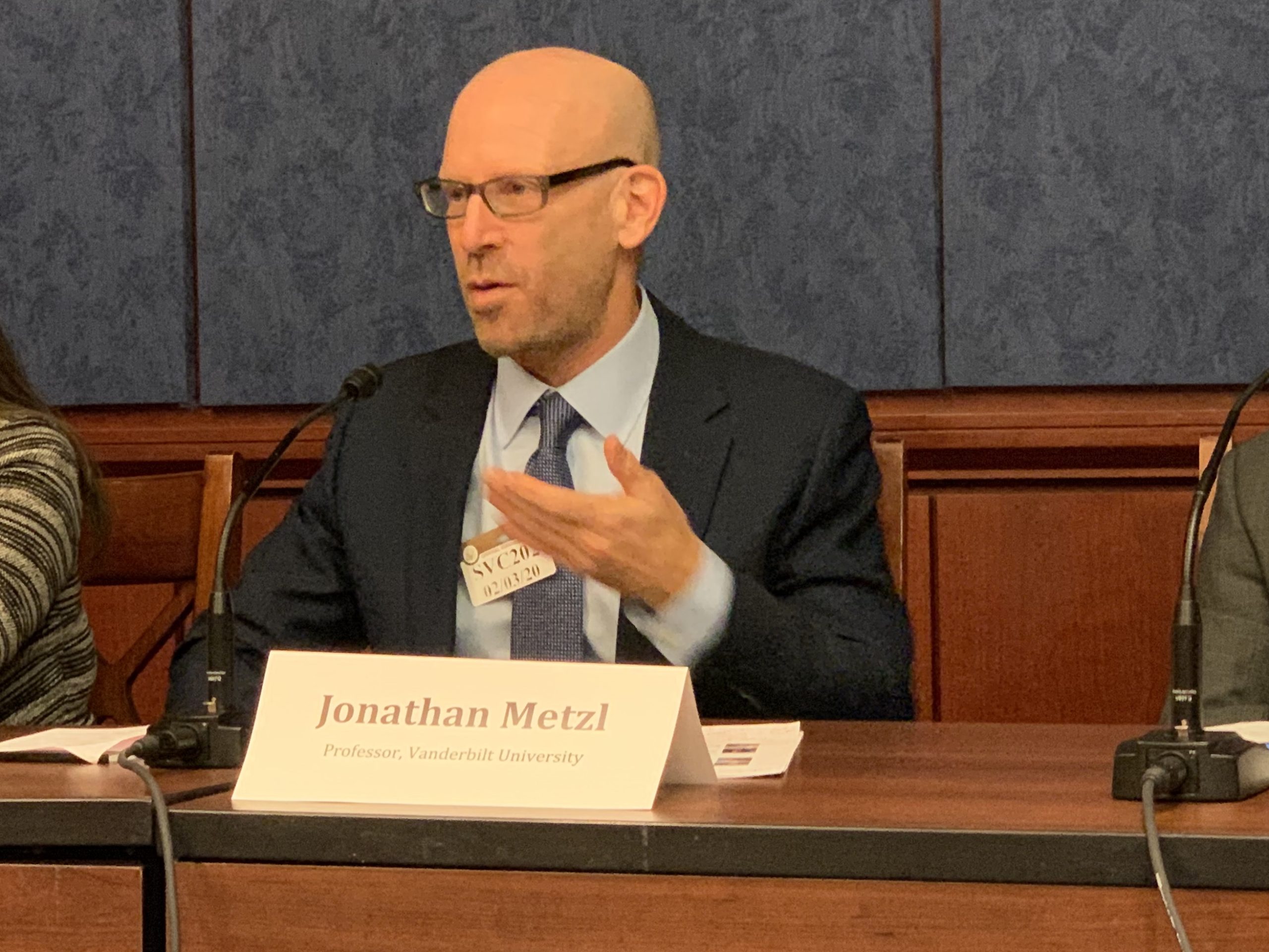 Jonathan Metzl wears a dark grew suit and speaks into a table microphone.