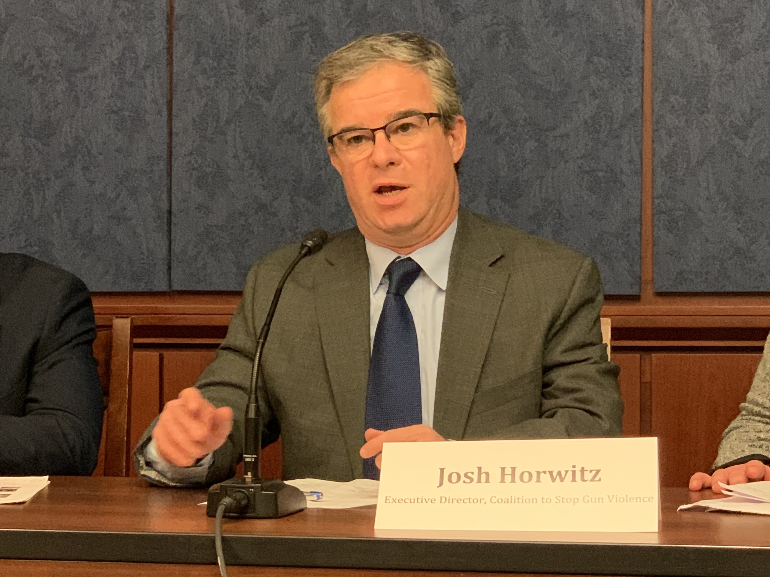 Josh Horwitz wears an olive suit and blue tie and speaks into a table microphone.