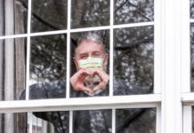 Facing the Coronavirus Crisis Together Could Lead to Positive Psychological Growth Man wearing a mask makes a heart with his hands from behind a window.