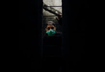 What COVID-19 Has to Teach Us About Psychiatric Oppression woman in medical mask peers out of darkness