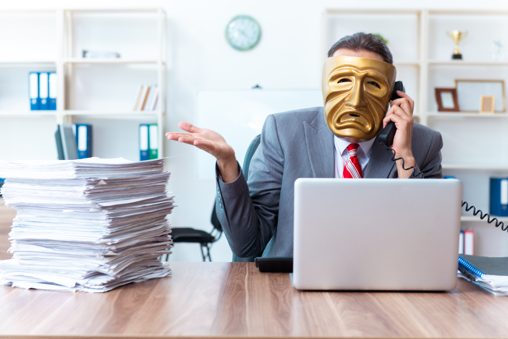 Photo of a man in a suit wearing a mask at a desk with a computer and a tall pile of paperwork