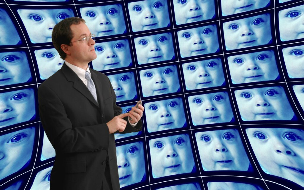 photo of man in suit standing in front of a wall of TVs with distorted babies' faces on them