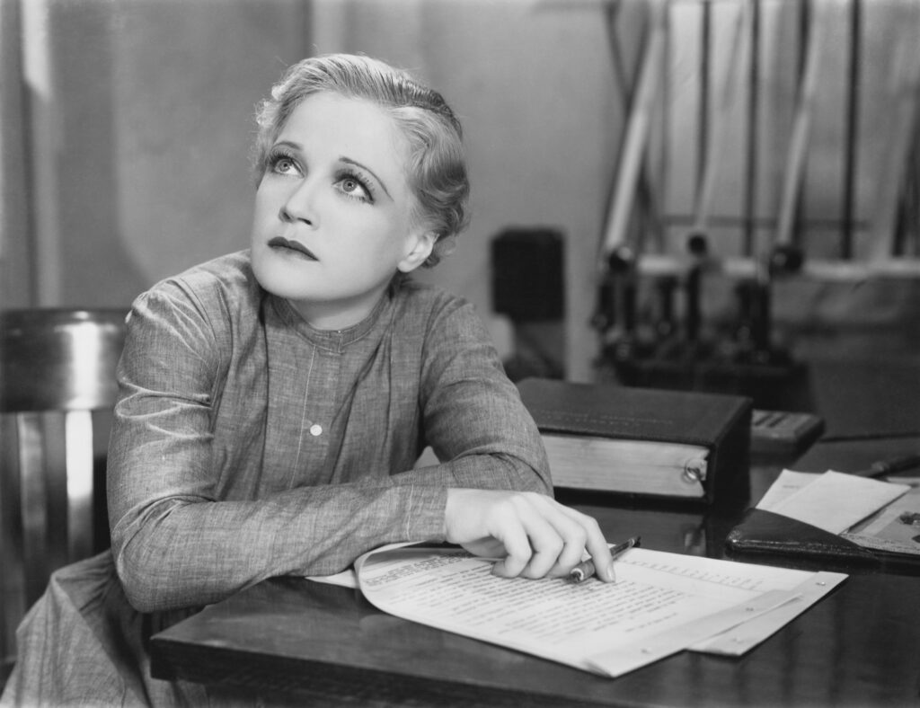 Black and white photograph depicting a seated woman looking up thoughtfully from a pile of papers on a desk