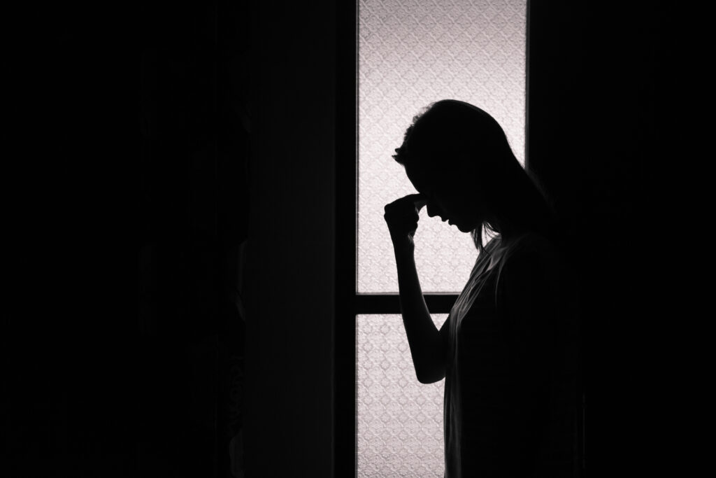 Photo of a person sihouetted against a window holding their face with their hand, looking sad or in pain