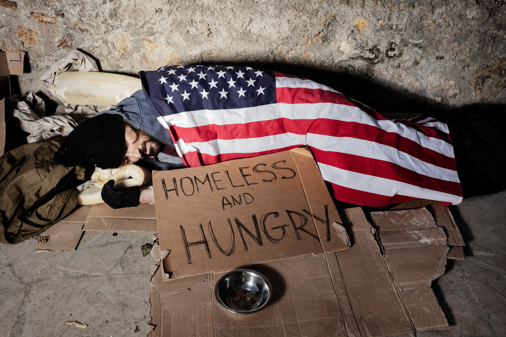 Homeless man begging for alms, sleeping on the sidewalk floor covered with American flag