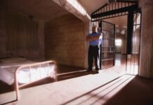 Inside a Forensic Psychiatry Unit: Earning the Right to Sleep on the Floor Photo of a prison cell with an uncomfortable-looking bed in the corner; a guard stands by the door