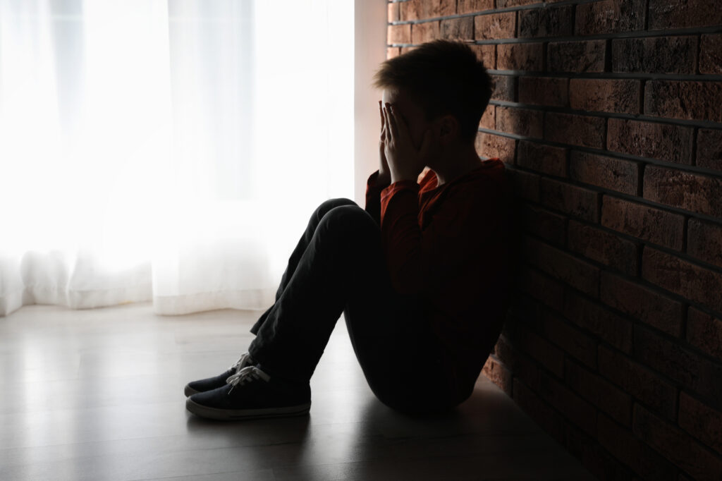 Photo of a boy sitting against a brick wall with his hands covering his face