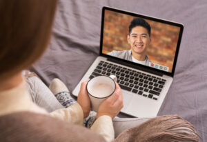 Photo shows back view of woman sitting holding a tea with a laptop computer with a young Asian man smiling on the screen