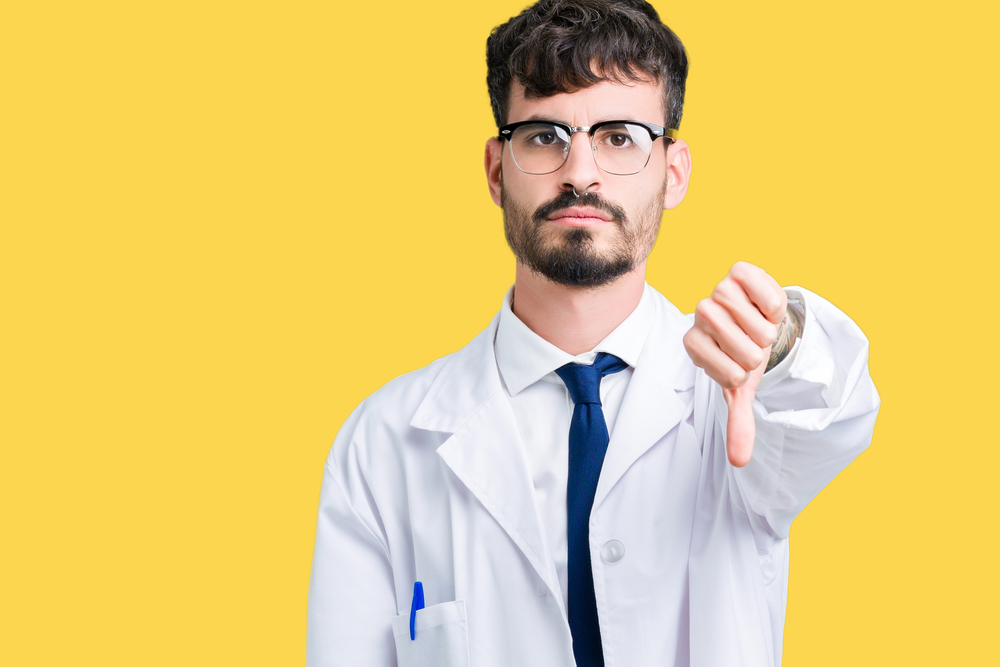 Young professional scientist man wearing white coat over isolated background looking unhappy and angry showing rejection and negative with thumbs down gesture. Bad expression.