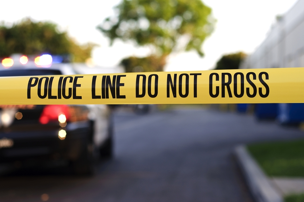 Crime scene tape in the foreground with a blurred police car in the background at a crime scene.