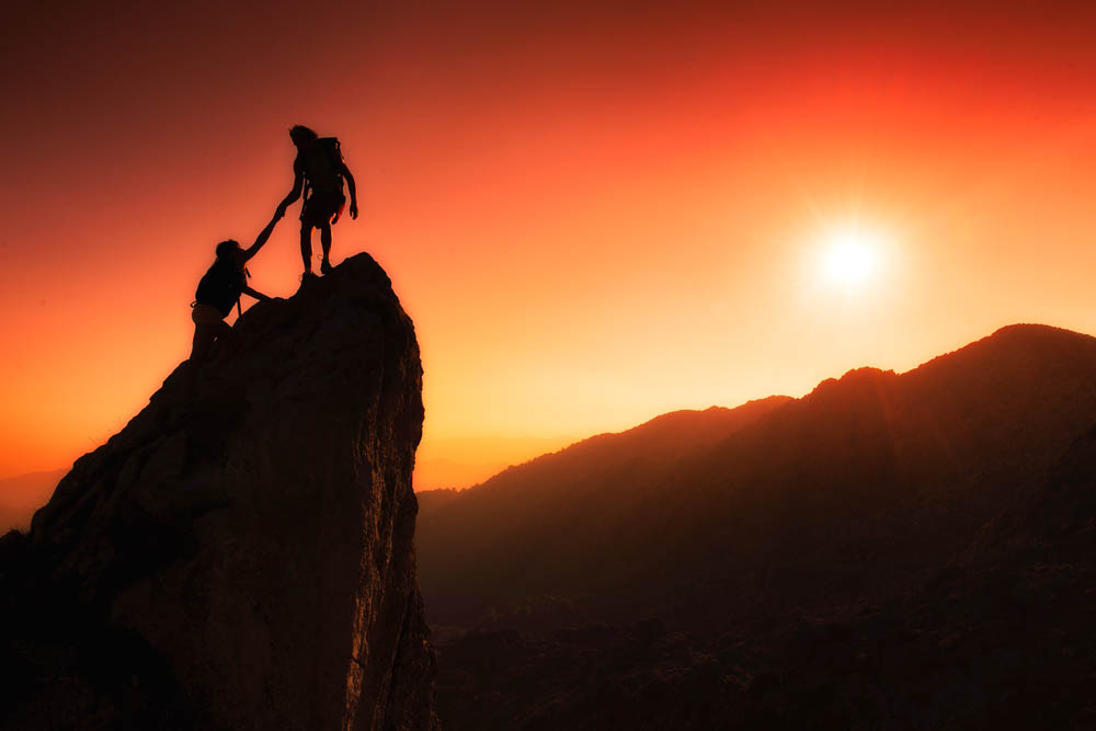 Photo of two silhouetted people on top of a rock at sunset. One is helping the other.