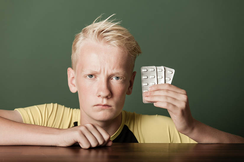 Scowling teenage boy holding up pills sealed in blister packs in his hand as he leans on a wooden table with an intense stare