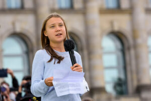 Greta Thunberg, young white Swedish woman, speaking into microphone in front of large building.