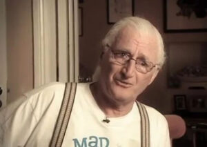 John McCarthy, white older Irish man with white hair and glasses in Mad Pride t-shirt and suspenders. In front of a wall with framed photos and a doorframe.