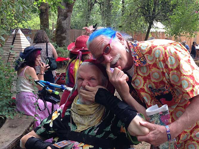 Blog author, David Oaks, in wheelchair with Patch Adams, with blue hair and glasses. Both are picking their noses at the Oregon Country Fair, with trees in the background. Oaks says, "Searching for meaning."