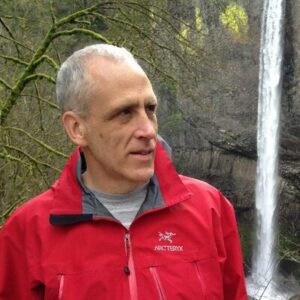 Ron Unger, an older white man in a red jacket in front of trees and a small waterfall.