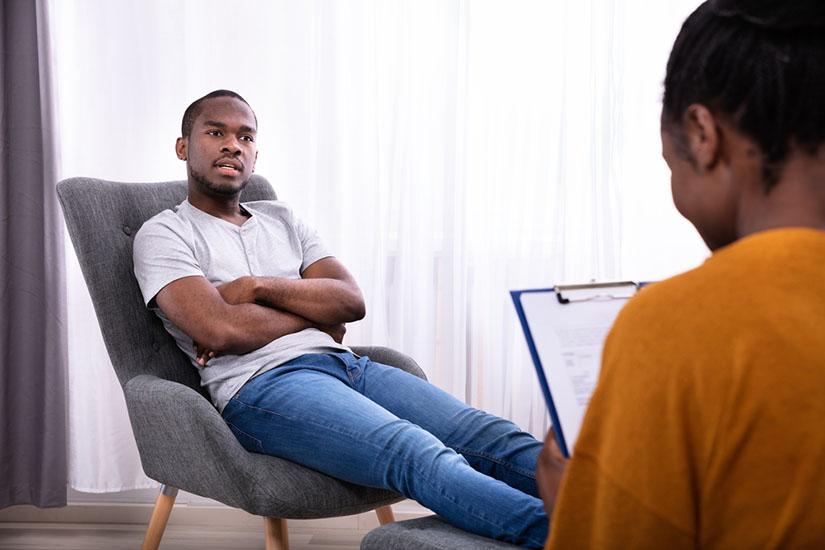 Young African Man Sitting On Chair Near Female Psychologist With Clipboard