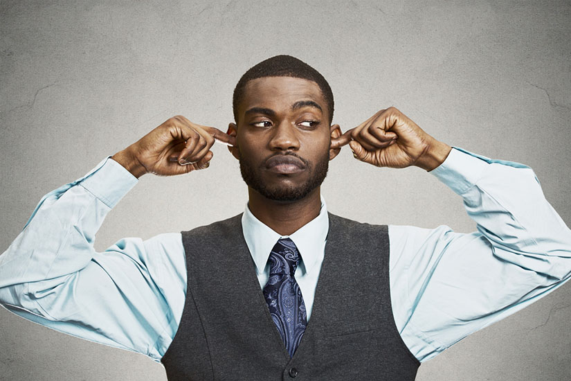 A black man in a suit stands with fingers in his ears as if pretending not to hear. His eyes to the side as if nervous.