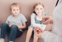 No Evidence for Long-Term Safety or Efficacy of Mental Health Treatment in Children A boy and a girl, about 5 years old, sit on a couch, slightly out of focus. On the right, two hands enter frame: one holds a tall glass full of water; the other holds a bunch of colorful pills.