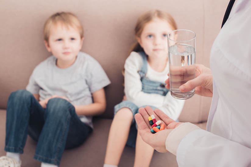 A boy and a girl, about 5 years old, sit on a couch, slightly out of focus. On the right, two hands enter frame: one holds a tall glass full of water; the other holds a bunch of colorful pills.