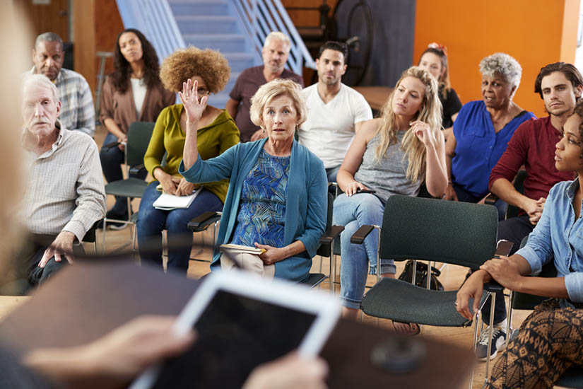 Seated among others at a town hall meeting, an older adult white woman raises her hand