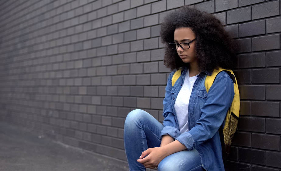 Young Black girl wearing a backpack sitting against a brick wall, looking sad