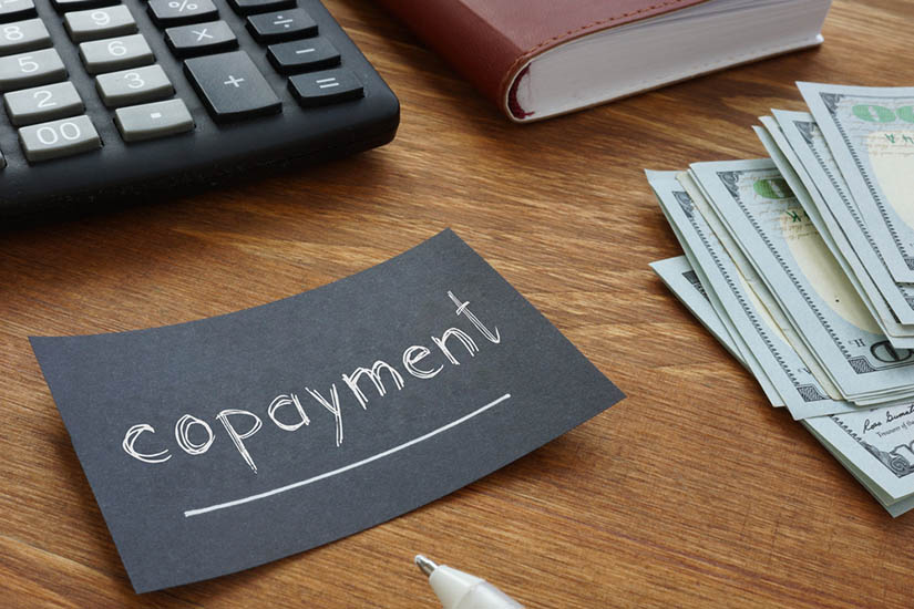 Close-up of the wood top of a desk. On it: money (bills), a calculator, a notebook, and prominently featured, a black business card reading in white text, underlined, "copayment."