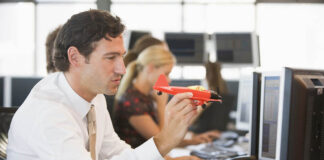 Meaningless Distractibility, or Meaningful Mind-Wandering? Photo of a man in a white button-down and tie, sitting at a desk in an office, with one hand on a computer keyboard and the other holding a red toy airplane