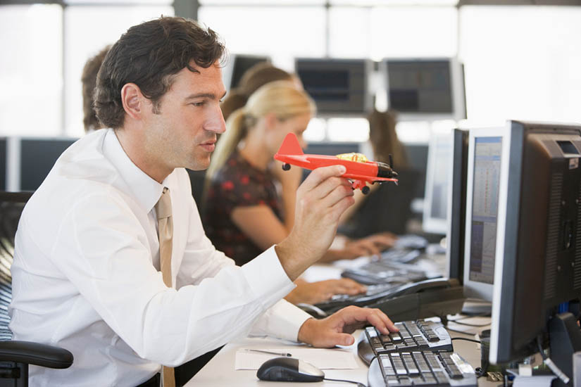 Photo of a man in a white button-down and tie, sitting at a desk in an office, with one hand on a computer keyboard and the other holding a red toy airplane