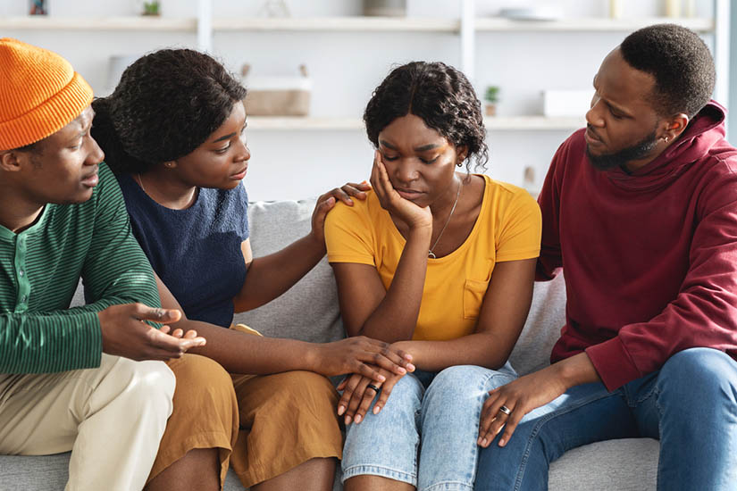 Four African-American young adults on a couch. In the center, a woman wearing yellow and looking sad is being comforted by a man and a woman on either side.