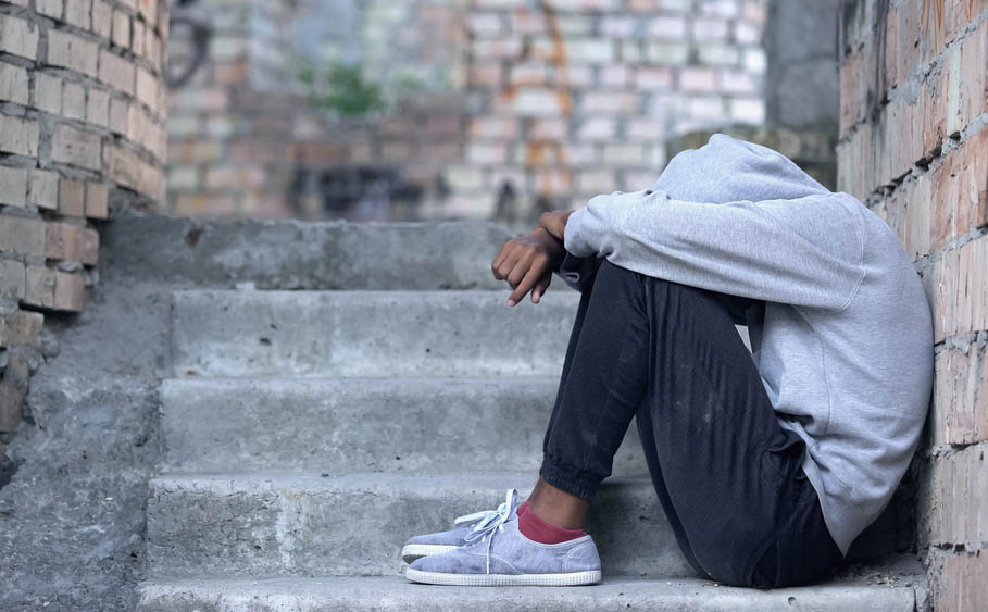 A black teen in a hooded sweatshirt sits on the stairs, hiding their face, looking depressed.