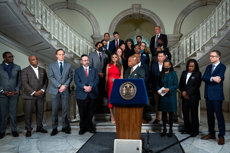 A photo of Mayor Eric Adams at a podium, with many other people behind him wearing suits.