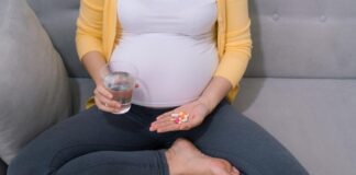 Dubious Science: Downplaying the Risks of Antidepressants in Pregnancy Photo of pregnant person holding a glass of water and a handful of pills. Sitting cross-legged on couch. We cannot see her face.