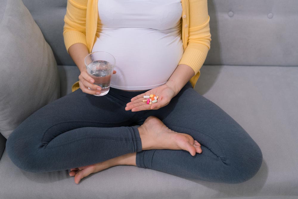 Photo of pregnant person holding a glass of water and a handful of pills. Sitting cross-legged on couch. We cannot see her face.