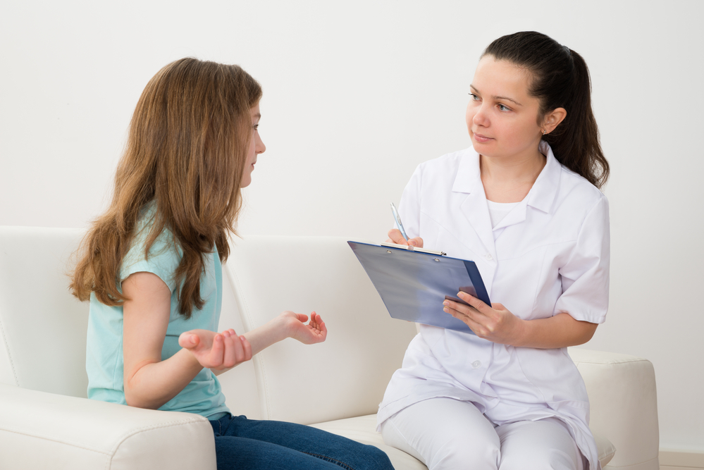 Female Doctor Writing On Clipboard With Patient Sitting On Sofa