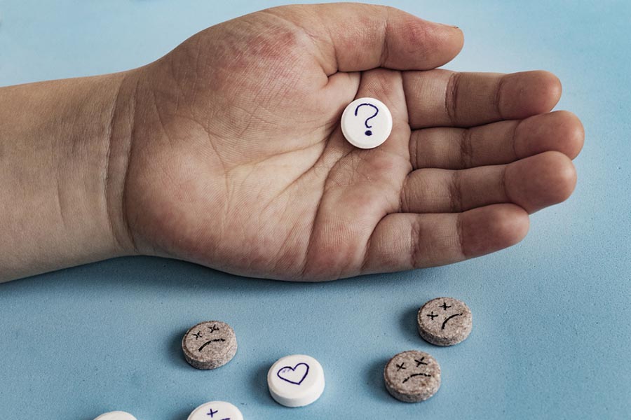 Close-up of a hand holding a tablet with a question mark on it. Below, scattered tablets with frowns or hearts.