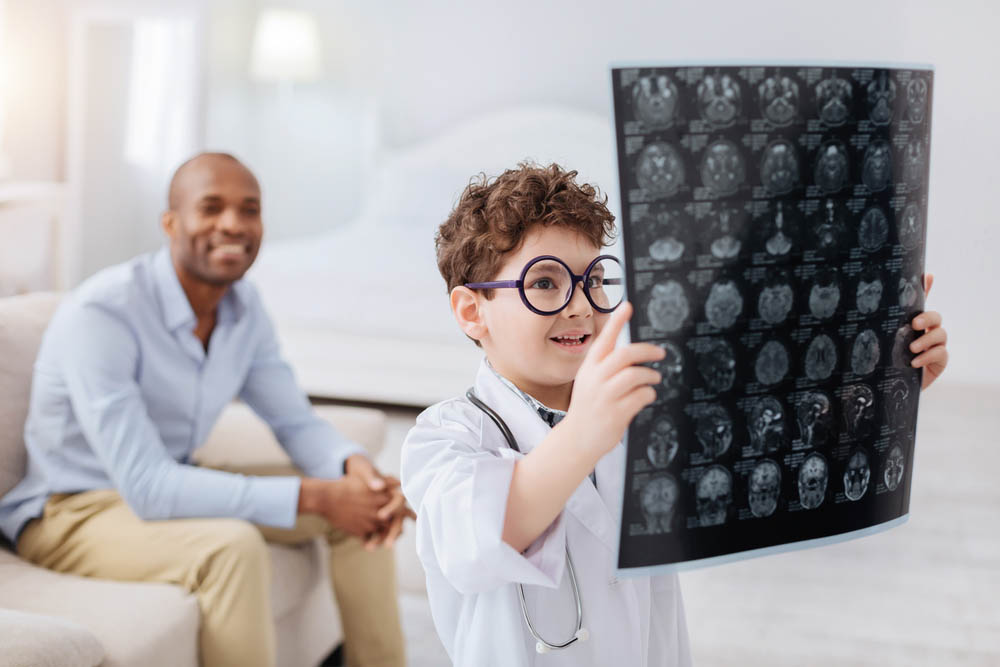 Photo of a young White boy with glasses looking at a giant sheet of brain imaging results, with a Black man smiling happily behind him