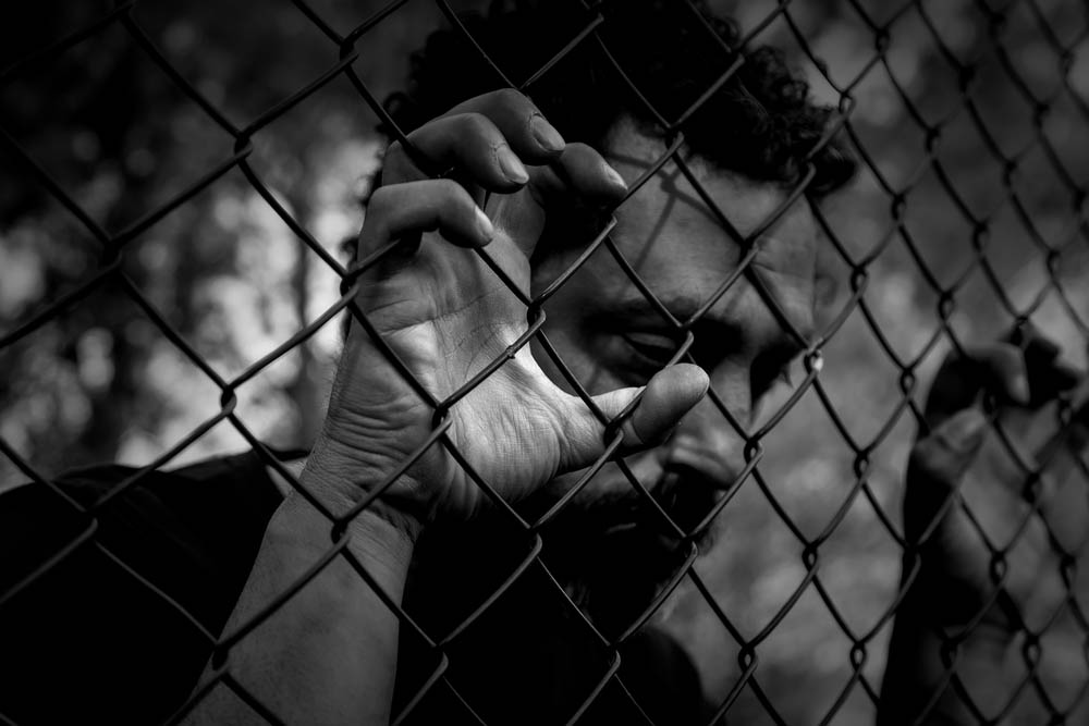 Black and white photo of a man looking sad with his fingers clinging to a chain-link fence