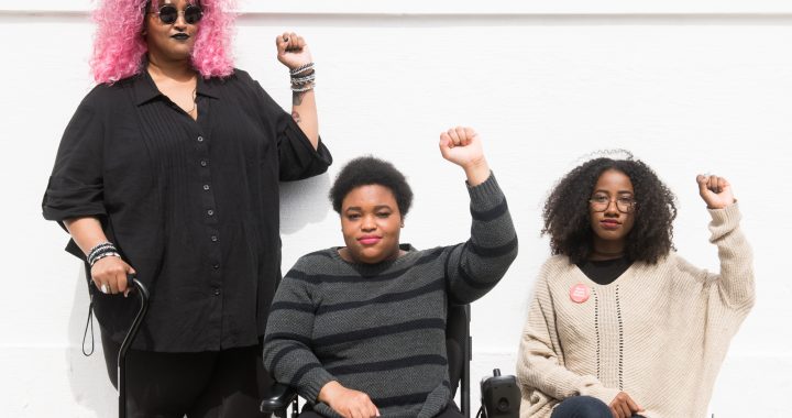 orso level photo of three Black and disabled folx (a non-binary person holding a cane, a woman in a power wheelchair, and a woman on a folding chair) raising their fists on the sidewalk in front of a white wall. (Credit: Disabled and Here.)