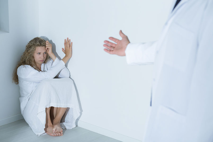 A young woman crouches in distress in a corner, while a doctor's hand reaches out to her. Everything is white