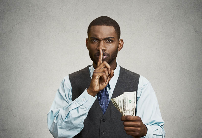 Photo of a man in a suit holding money and making a shush sign with his finger on his lips