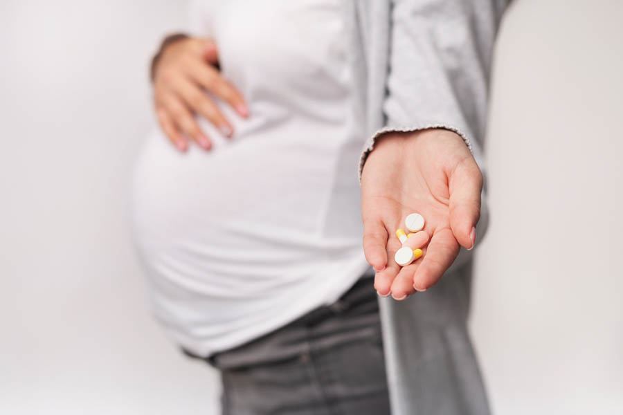 Photo of a pregnant person's belly clothed in jeans and shirt, hand holding out some pills