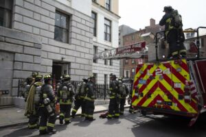 Several fire fighters in full gear stand outside the state house with the end of a fire truck visible behind them, and the fire truck ladder going into one of the building's windows