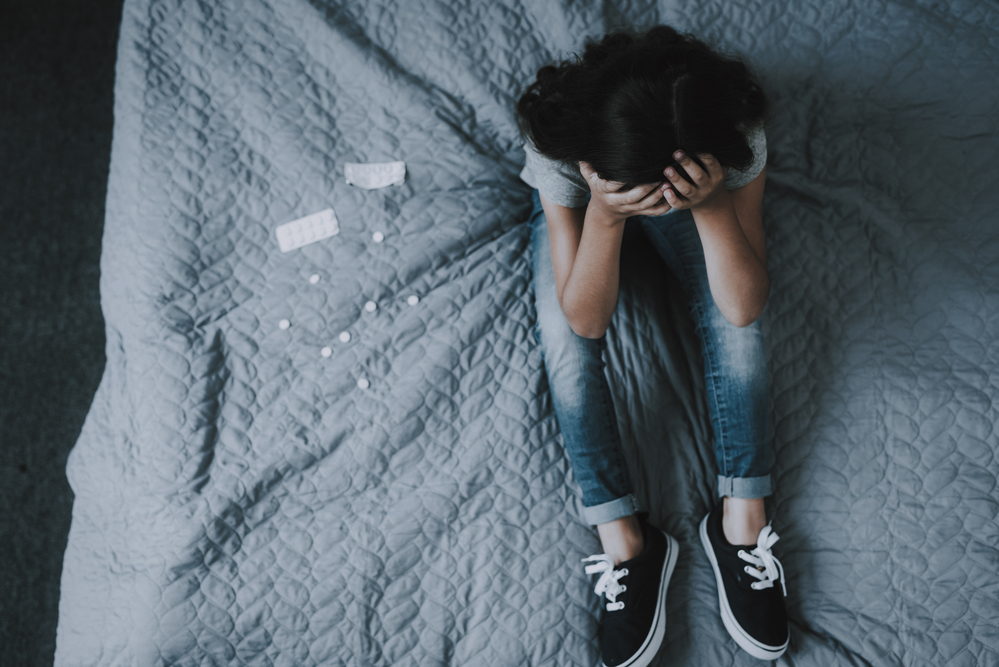 Top View of Girl Sits on Bed with Scattered Pills. Little Child Sitting on Gray Badcover Near Opened Packing of Medicines Keeps Head with Hands. Unhappy Childhood Concept