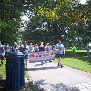 Several people wearing blue 'Mad Pride' shirts are visible walking down a wide, paved pathway. Two people in front are carrying a purple 'Vermont Mad Pride' banner. Grass and trees are visible in the background, and a trashcan is visible in the left foreground.