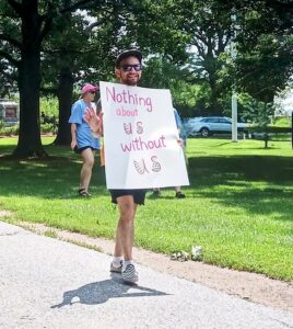 Male, light skinned individual with a beard and mustache, wearing sunglasses and a hat, shorts and a t-shirt is visible looking toward the camera, smiling and waving with his right hand while carrying a sign that reads "Nothing about us without us." A couple of individuals are visible in the background behind him, as is grass and trees.